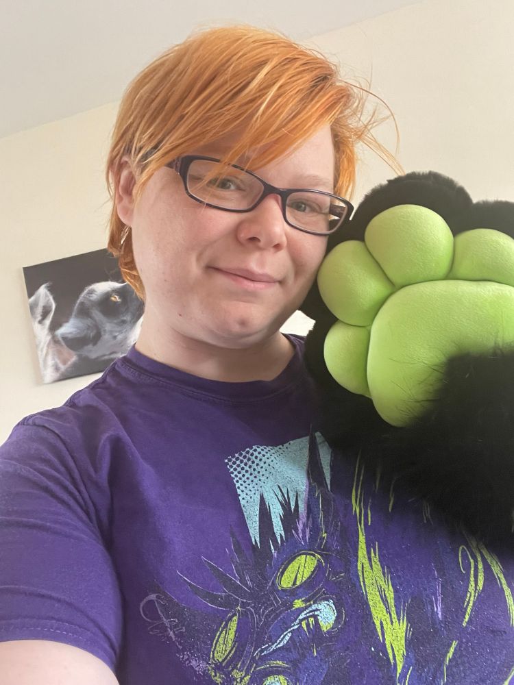 A trans masculine person wearing a purple shirt with glasses and orange hair takes a selfie showing off a large fursuit handpaw. The handpaw is all black, showing all lime green squishy beans. The paws are a bappy style with no individual fingers. 