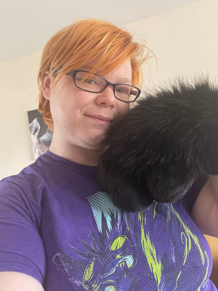  trans masculine person wearing a purple shirt with glasses and orange hair takes a selfie showing off a large fursuit handpaw. The handpaw is all black, showing the luxurious fur on the back . The paws are a bappy style with no individual fingers. 