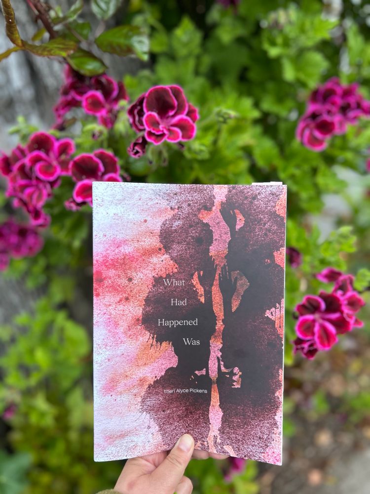 a hand holds up a copy of What Had Happened Was by Theri Pickens against a plant with dark pink flowers. The cover is various shades of pink, with a brown, ink-blot like repeating pattern of women whispering to each other. 