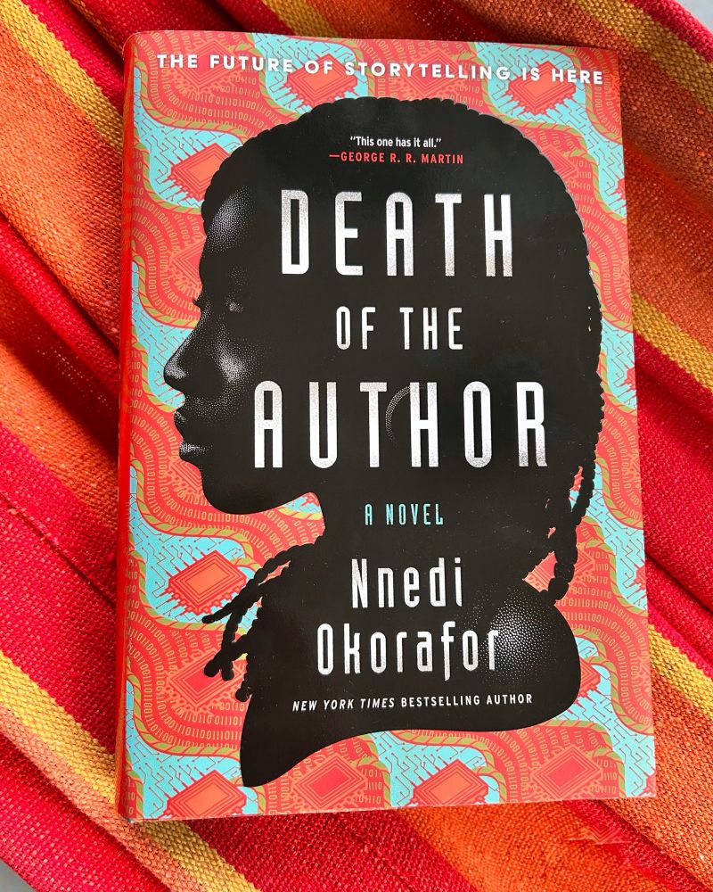 the book Death of the Author by Nnedi Okorafor sits on top of a red and orange hammock. The book cover has a red and blue Ankara-like pattern with squares that look like piece of technology. The silhouette of a Black woman’s face looking to the left is at the center. She has braids and the title text is white down the center of her head