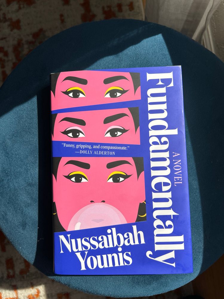 The book Fundamentally by Nussaibah Younis is pictured on a blue footstool. The book has a blue background and white title text with three spliced images of a pink feminine face. The bottom image of the face is blowing a bubble with gum.