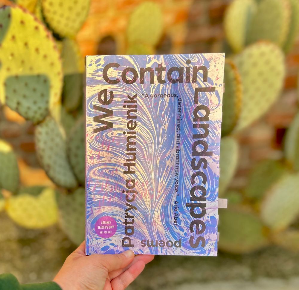 a hand holds up a book in front of a large cactus. The book, We Contain Landscapes, has black title text and a blue, white and purple swirling design on the cover.