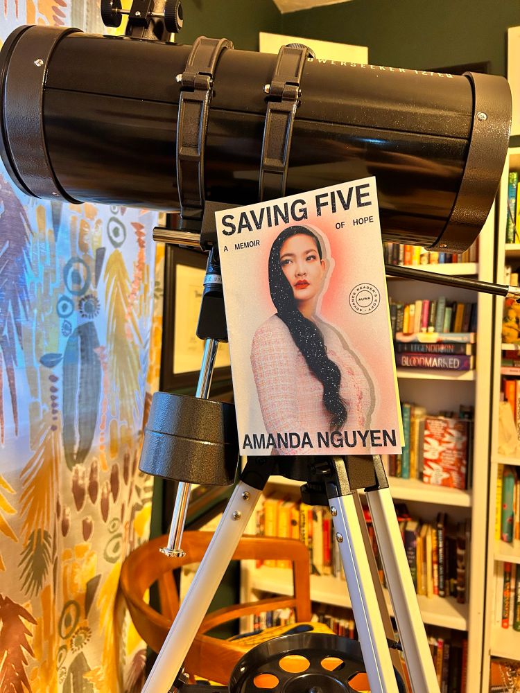 The book Saving Five by Amanda Nguyen is propped up against a large black telescope. The book cover has a portrait of the author, a Vietnamese woman with long black hair wearing a pink sweater. There is a shadow effect behind her and stars overlaid on her hair. 