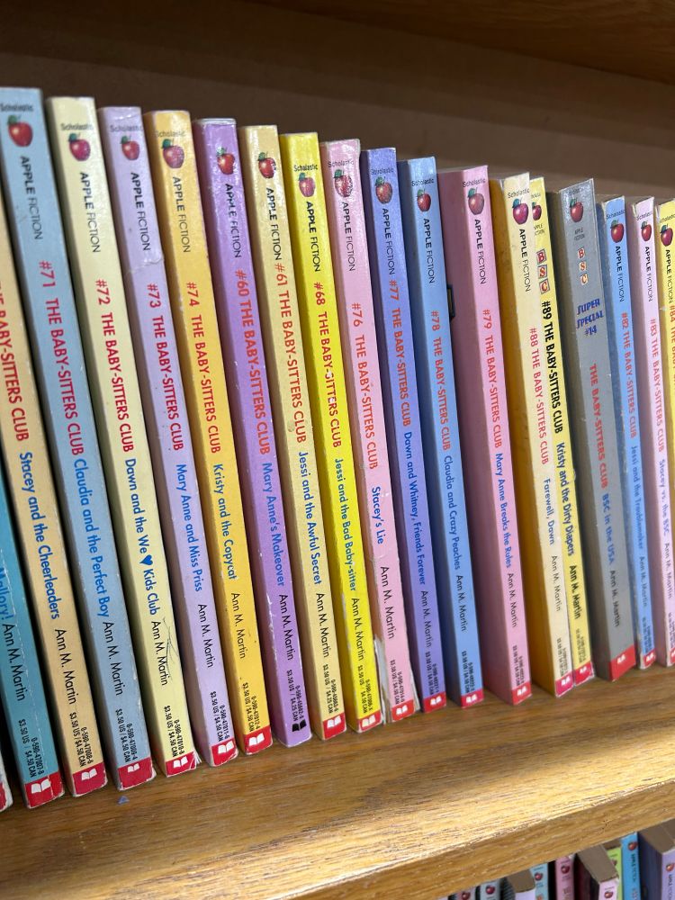 On top of a wooden shelf is a series of babysitters club books. 