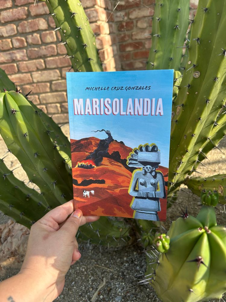 The book Marisolandia is held in front of a cactus. The book cover has a barren landscape and blue sky, with a stone statue in the bottom right corner