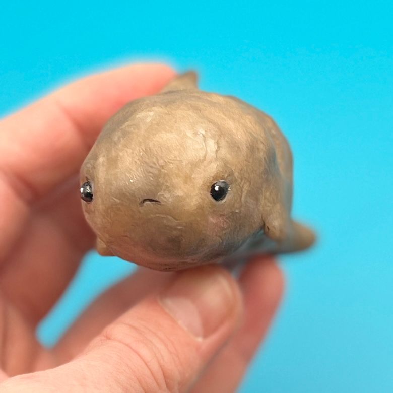 Against a solid background, a hand holds a small sculpture of a potato whale hybrid with a cartoonish sad facial expression