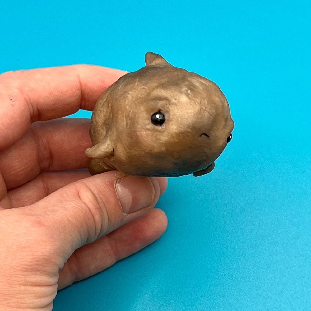 Against a solid background, a hand holds a small sculpture of a potato whale hybrid with a cartoonish sad facial expression