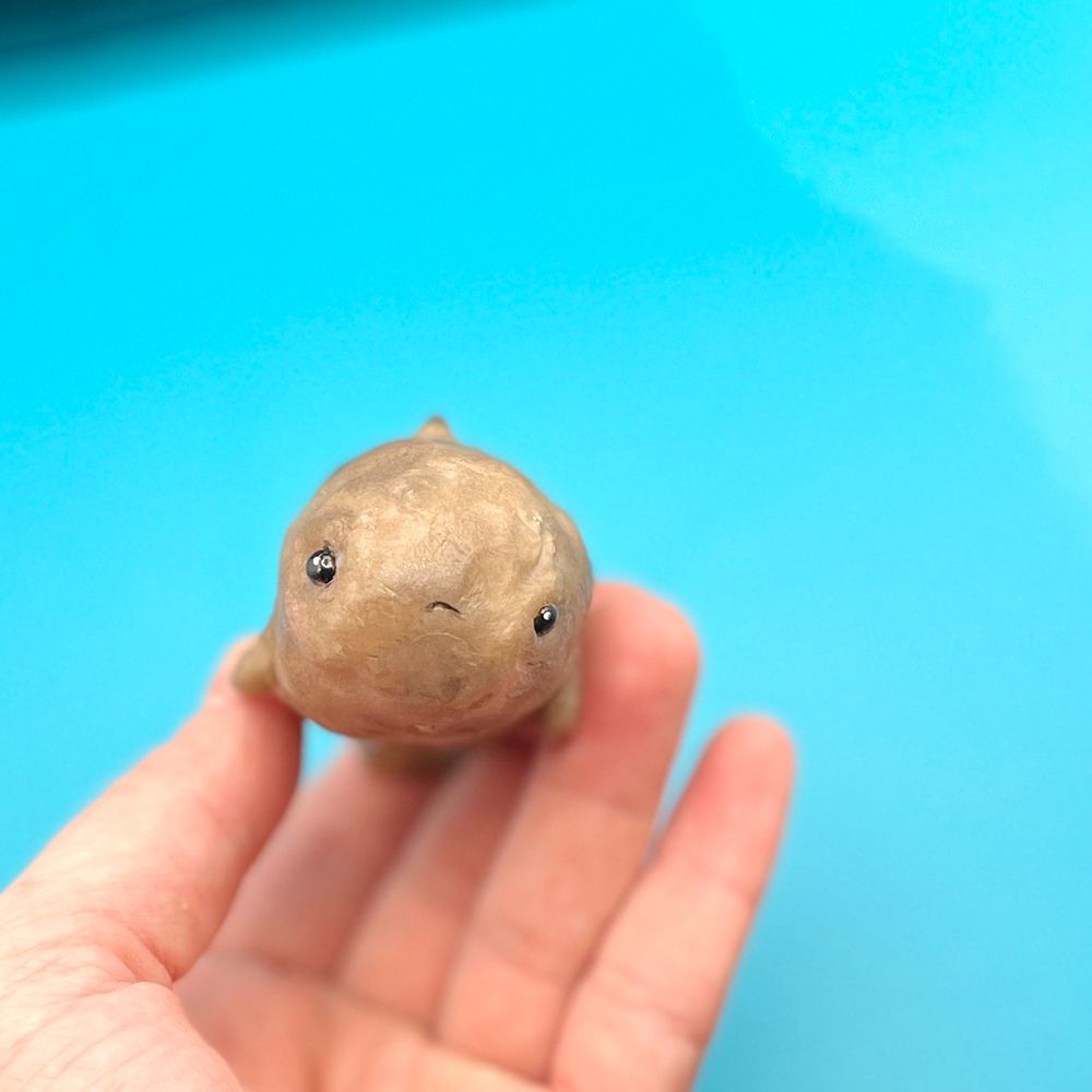 Against a solid background, a hand holds a small sculpture of a potato whale hybrid with a cartoonish sad facial expression