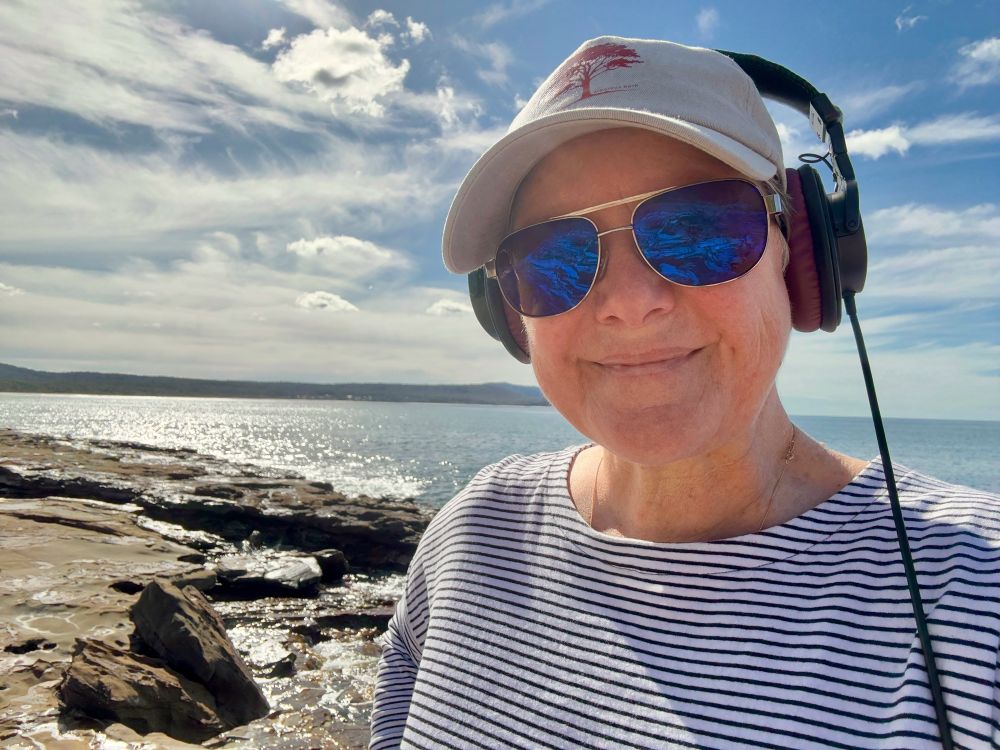 Me wearing a striped tee, sunglasses, a cap and headphones standing on a rock ledge by the ocean.