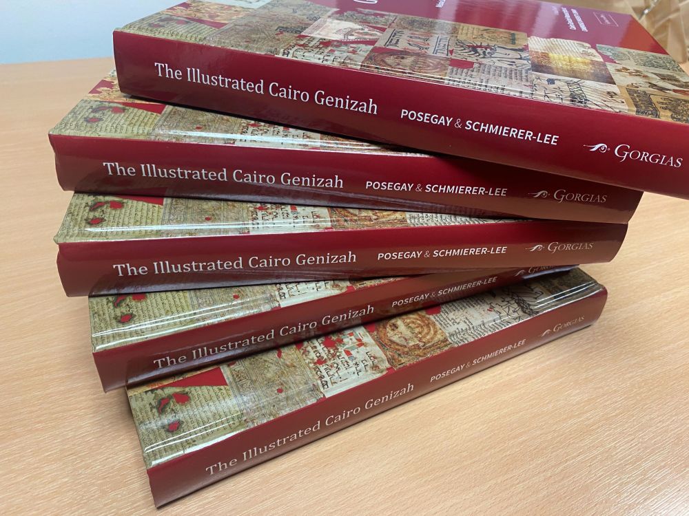 5 copies of 'The Illustrated Cairo Genizah', by Nick Posegay and Melonie Schmierer-Lee, stacked on a light brown tabletop. The books' spines are facing the camera. The book is large and the covers are dark red, with the title and authors' names in white. The cover also has a grid of 21 square images each cut out from a different medieval manuscript.