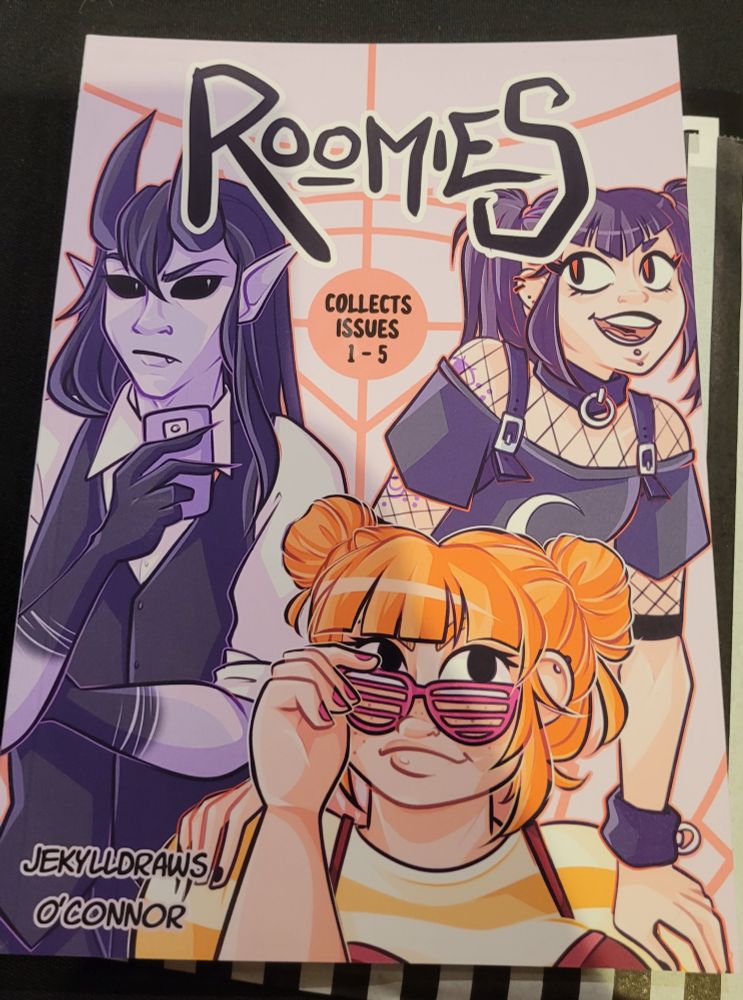 The front cover of the comic 'Roomies' showing 3 people: a long haired, black eyed, demon looking person with purple skin; a person with dark hair in bunches and goth and fetish clothing; and a person with orange hair in puff bunches and an orange striped shirt.