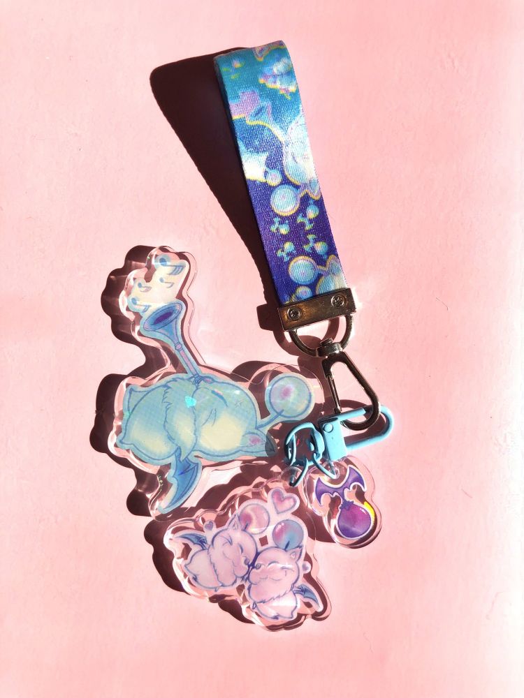A photo of a wrist lanyward featuring a moogle motif, with one blowing a horn, two nosing each other cutely with a heart above it, and one kupo nut. These illustrations are also on accompanying acrylic charms attached to the lanyard with a blue clip. The main color scheme is pastel blue, pastel pink, and some dark blue/indigo.