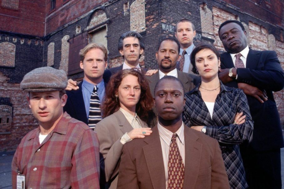 Characters from 90s police procedural show Homicide: Life on the Streets. 9 hardboiled Baltimore detectives, posed in stepped ranks in front of an abandoned looking brick warehouse or industrial building.