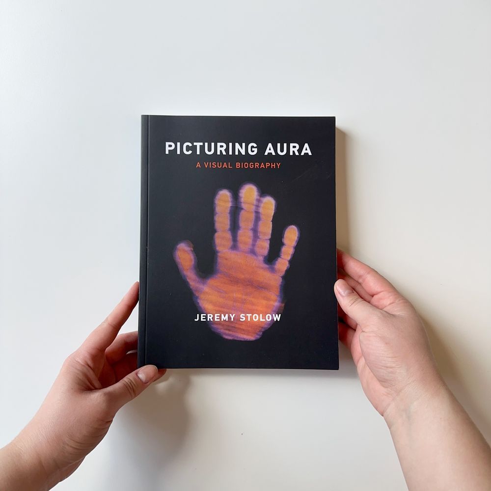 Hands holding a copy of "Picturing Aura: A Visual Biography" on a plain background. the cover is black with a red imprint of a hand.