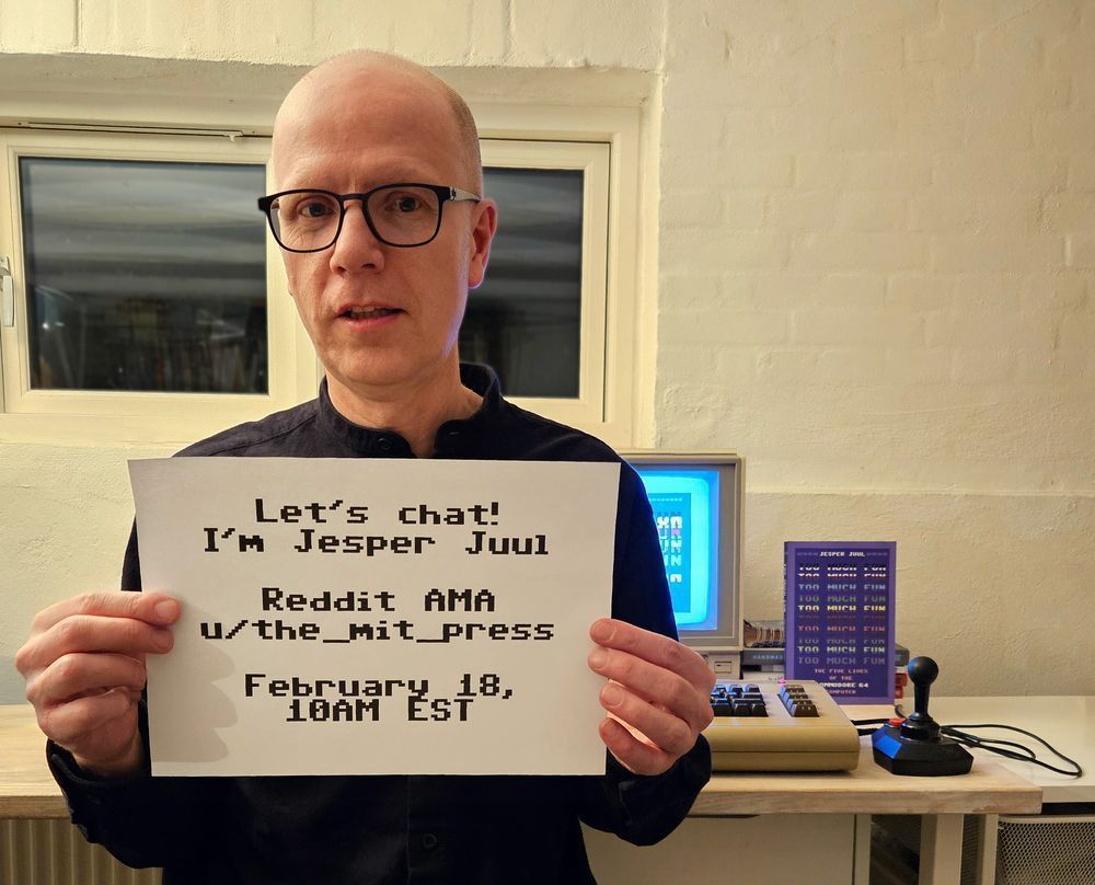 Photo of Jesper Juul holding a piece of paper that reads "Let's chat! I'm Jesper Juul Reddit AMA u/the_mit_press February 18, 10 am EST" A copy of his book "Too Much Fun" is visible in the background next to a joystick.