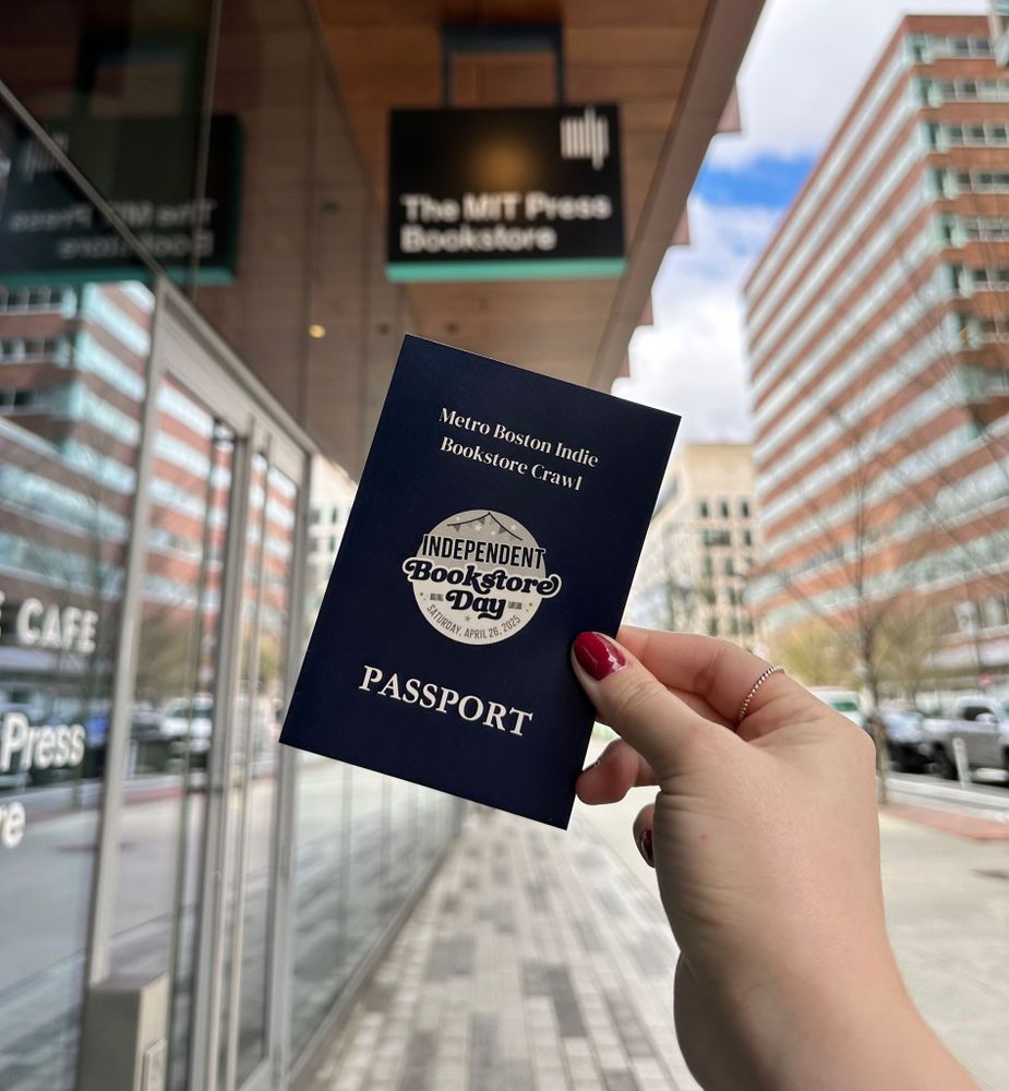 Hand holding a blue booklet outside a store front. The booklet is labeled "passport" and the logo Independent Bookstore Day is at the center. The top reads "Metro Boston Indie Bookstore Crawl." A sign for the MIT Press Bookstore is visible in the background.