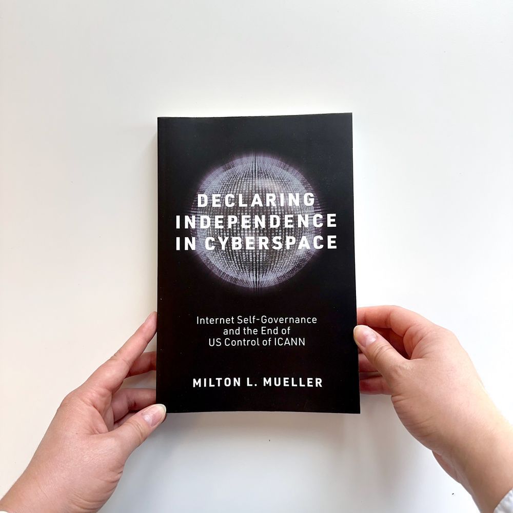 Hands holding a book entitled "Declaring Independence in Cyberspace: Internet Self-Governance and the End of US Control of ICANN" by Milton L. Mueller. At the center of the cover is a glowing orb comprised of binary numbers.