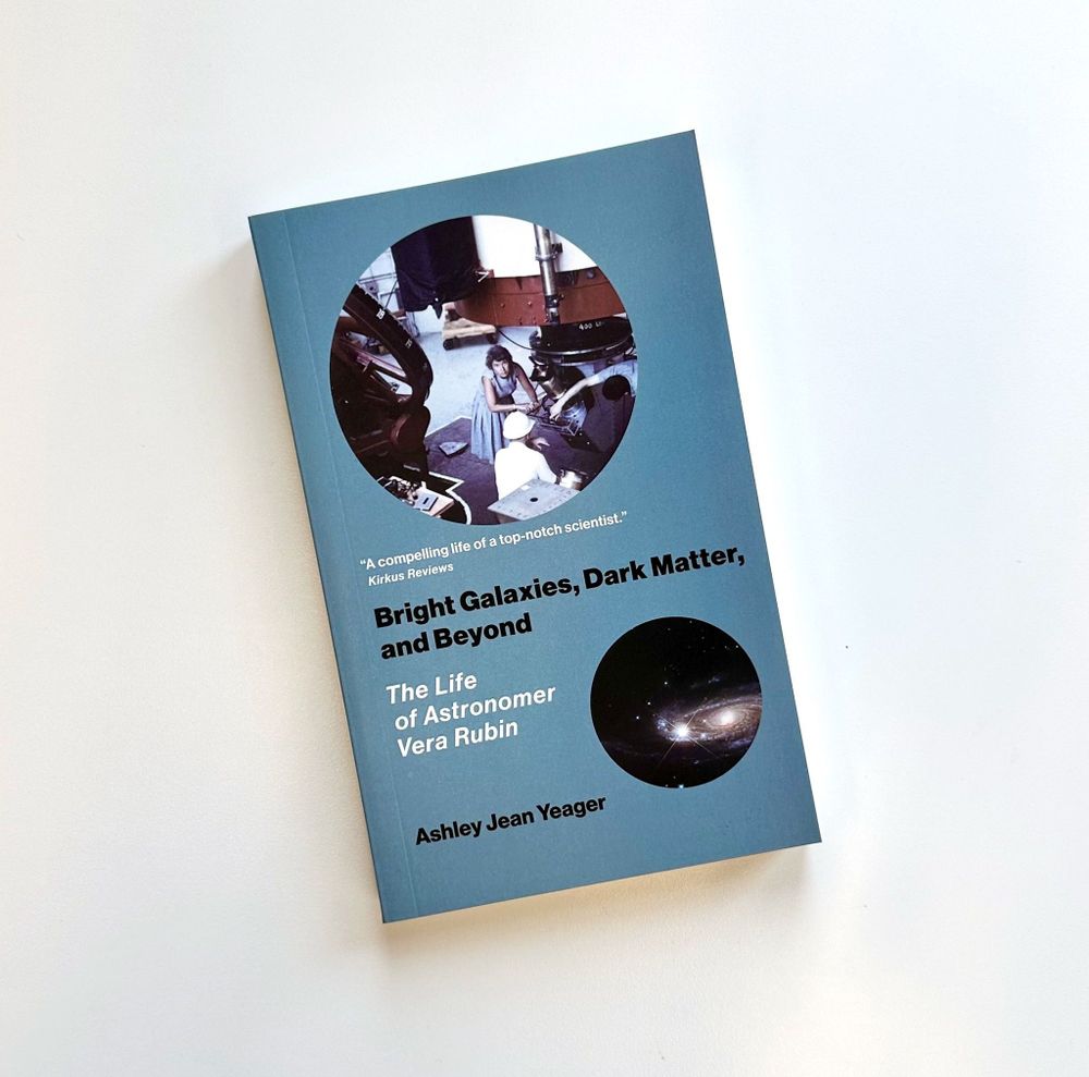 Copy of "Bright Galaxies, Dark Matter, and Beyond: The Life of Astronomer Vera Rubin" by Ashley Jean Yeager on a white background. The cover is blue with a picture of Rubin working on a telescope, and a photograph of a galaxy. 