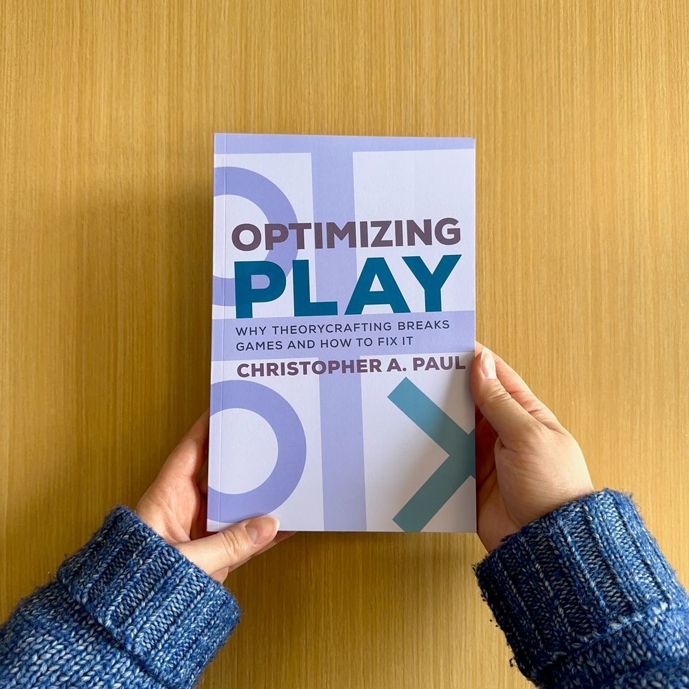A person holding a book titled "Optimizing Play: Why Theorycrafting Breaks Games and How to Fix It" by Christopher A. Paul, with a blue and white cover.