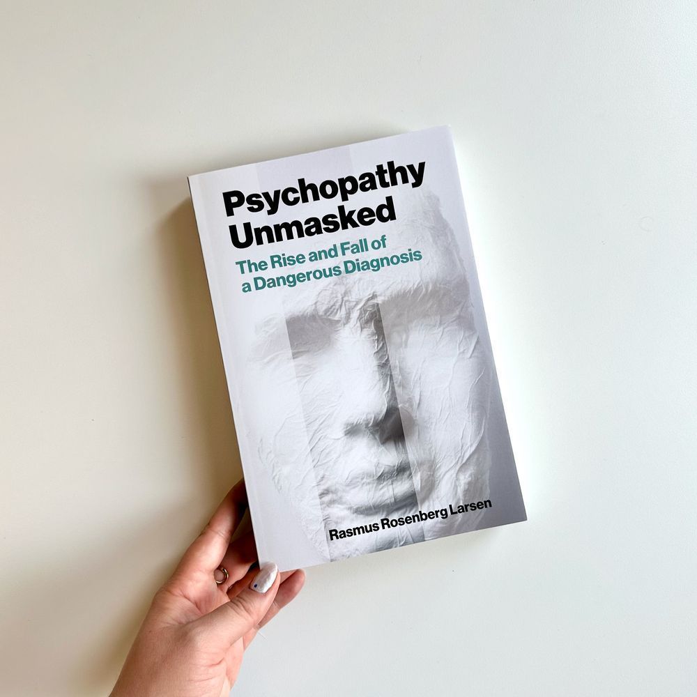Hand holding a copy of "Psychopathy Unmasked: The Rise and Fall of a Dangerous Diagnosis" by Rasmus Rosenberg Larsen. The cover is white and feature the front-facing profile of a white, paper mache mask.