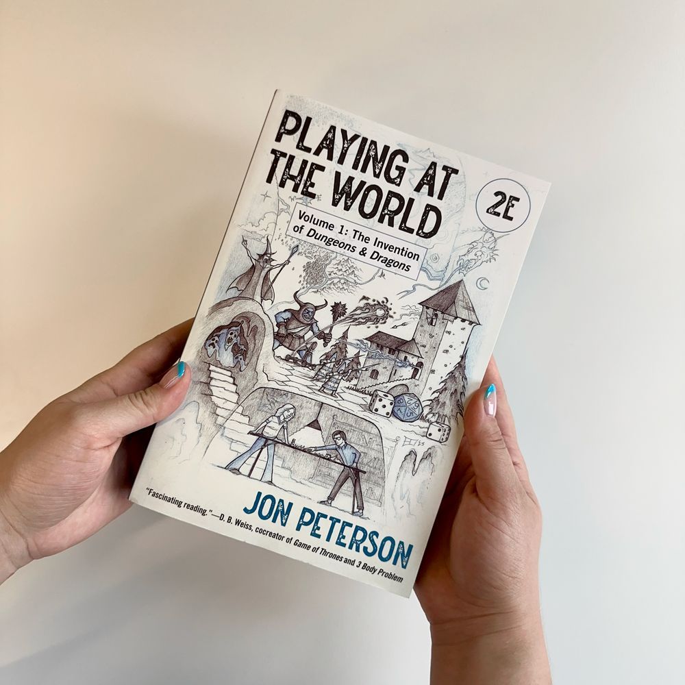 Hands holding a copy of Jon Peterson's "Playing at the World, 2E Volume 1: The Invention of Dungeons and Dragons." Illustrations of medieval characters and mythical monsters are depicted along the top half of the cover. Along the bottom half is an illustration of two figures are playing a board game under a hanging light.