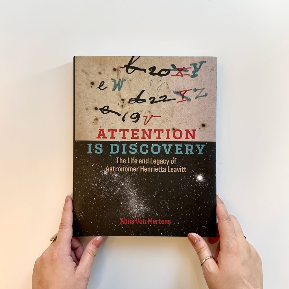 Hands holding a copy of "Attention is Discovery: The Life and Legacy of Astronomer Henrietta Leavitt" by Anna Von Mertens. The top half of the cover features notations from Leavitt's notes. The bottom half features a photograph of the stars.