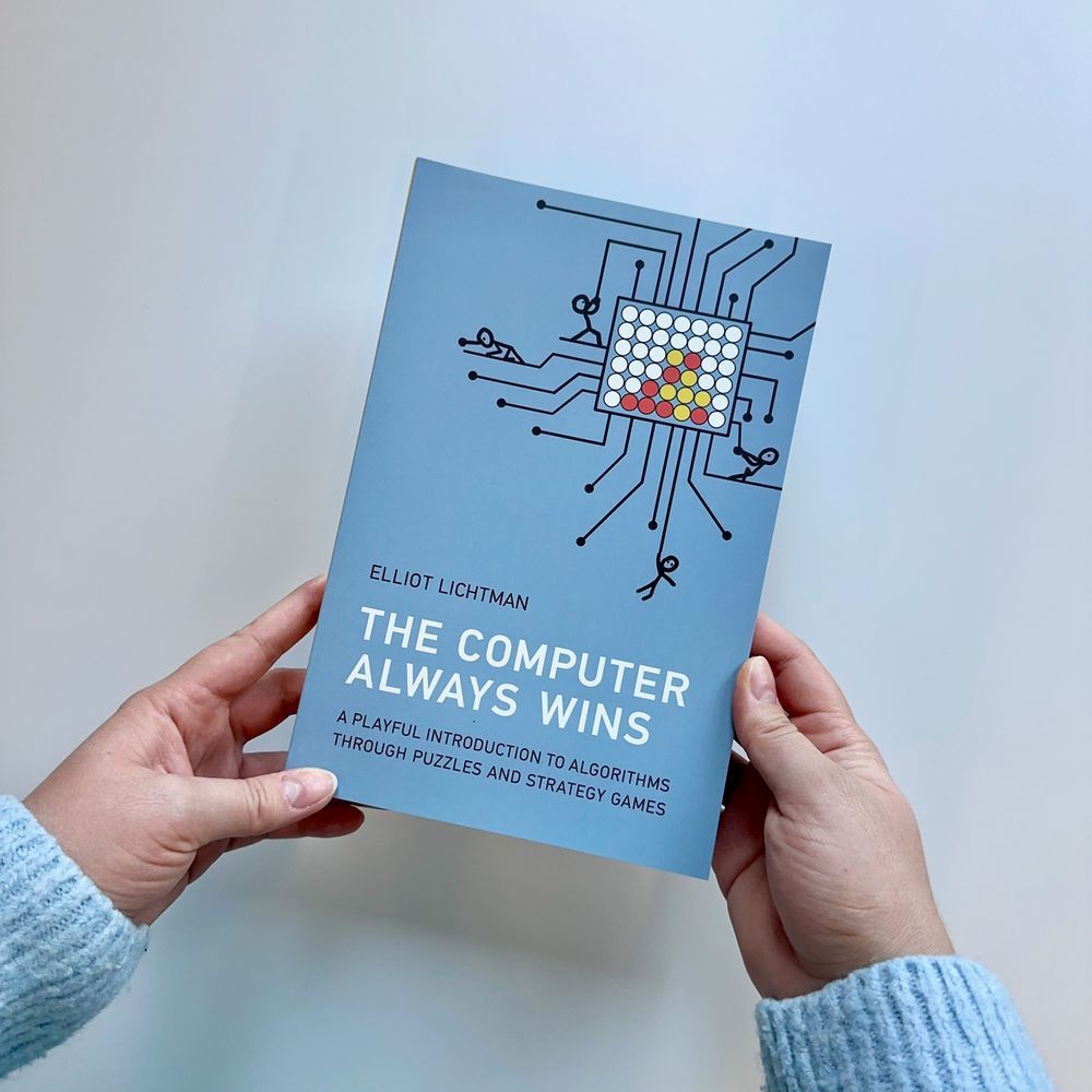 Hands holding a copy of "The Computer Always Wins: A Playful Introduction to Algorithms through Puzzles and Strategy Games" by Elliot Lichtman. The right corner of the cover features an illustration of a game of Connect 4 with stick men pulling on lines protruding from the board.