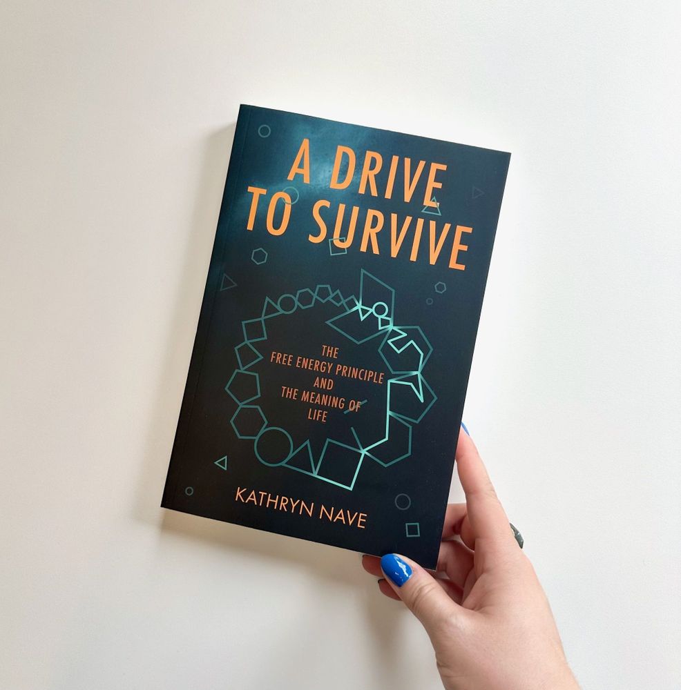 Hand holding a copy of "A Drive to Survive: The Free Energy Principle and the Meaning of Life" by Kathryn Nave. The cover is blue with orange text and features a geometric design at the center.