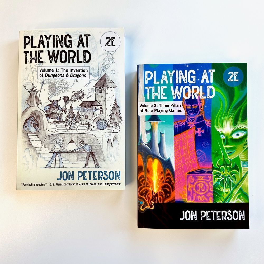 Copies of "Playing at the World, 2E Volume 1: The Invention of Dungeons & Dragons," and "Playing at the World, 2E Volume 2: Three Pillars of Role-Playing Games" on a plain background. The covers feature illustrations of fantasy and medieval characters.