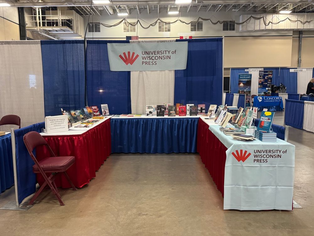 A photo of the University of Wisconsin Press booth at FinnFest 2025 in Duluth, MN, with three tables forming a U shape, with books set up on display all around. In back, the University of Wisconsin Press banner hanging atop blue and white curtains