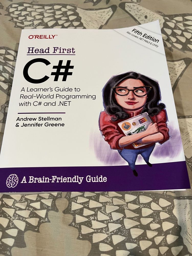 A tome of arcane knowledge rests upon a battlefield of shadows & light – a quilted tapestry where countless hedgehog sentinels stand eternal watch, their quilted forms echoing across the fabric like soldiers in an endless parade. This mystical backdrop, wrought in shades of storm-cloud grey & pearl, creates a fitting stage for the grimoire that commands centre stage – the legendary "Head First C#, Fifth Edition".


The shimmers with promise, its pristine white surface marked with the sigil of O'Reilly, blazoned in the colour of fresh-spilled wine. At its heart stands the rune "C#", bold as a warrior's declaration, flanked by the sacred inscription "A Learner's Guide to Real-World Programming with C# and .NET". The scribes who penned this codex – Andrew Stellman and Jennifer Greene – have etched their names beneath, testament to their mastery of the digital arts.


An illustrated guardian brings this book to life, as they stand watch upon its surface – an enchanting figure with raven-dark waves of hair & eyes that sparkle with wisdom behind crystalline spectacles. Clad in a garment the colour of autumn leaves, she cradles a laptop adorned with mystical sigils, her face in awe of the secrets yet untold. She is both teacher & guide, keeper of coding mysteries, ready to lead worthy students through the labyrinth of programming knowledge.