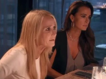 Two women at a table. One has blond hair and a white shirt and it bent forward. The other is brunette and wearing a black sweater and gray shirt