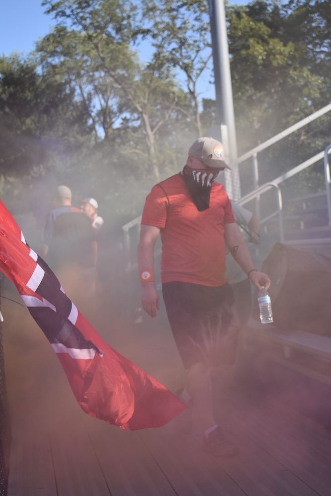 A white man, wearing a red shirt, a bandana, and dark shorts, is walking through smoke. In the background are outdoor bleachers and a sports team flag.