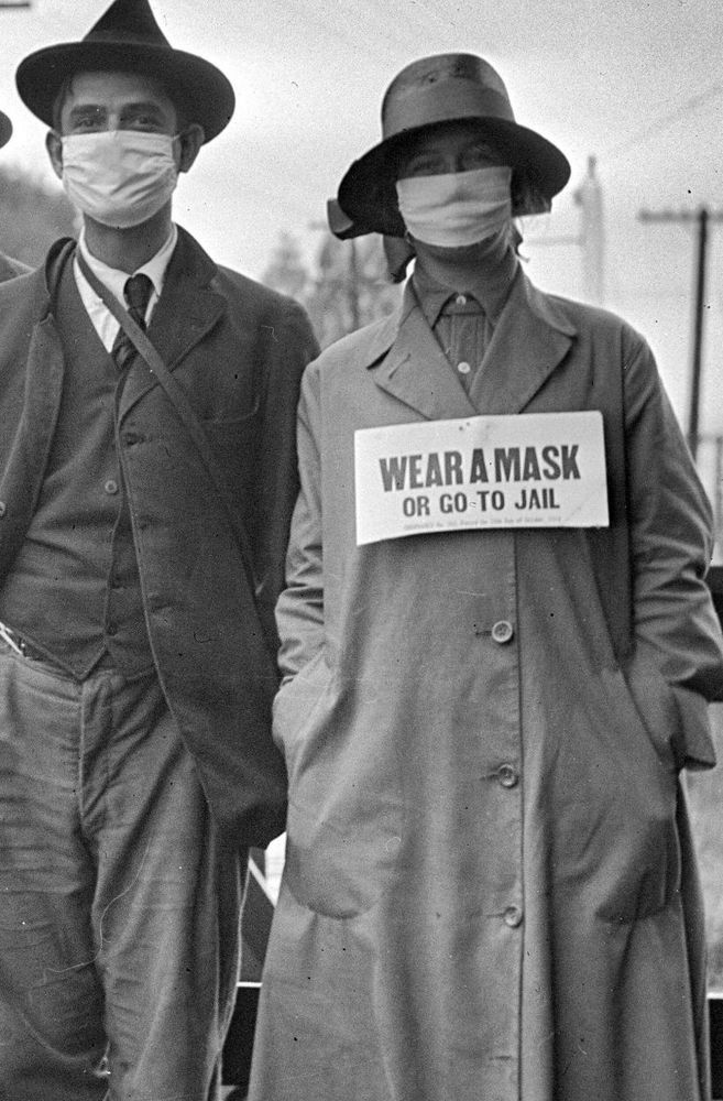 A man and woman wearing masks during the "Spanish Flu" epidemic.  She also wears a sigh saying "WEAR A MASK OR GO TO JAIL".