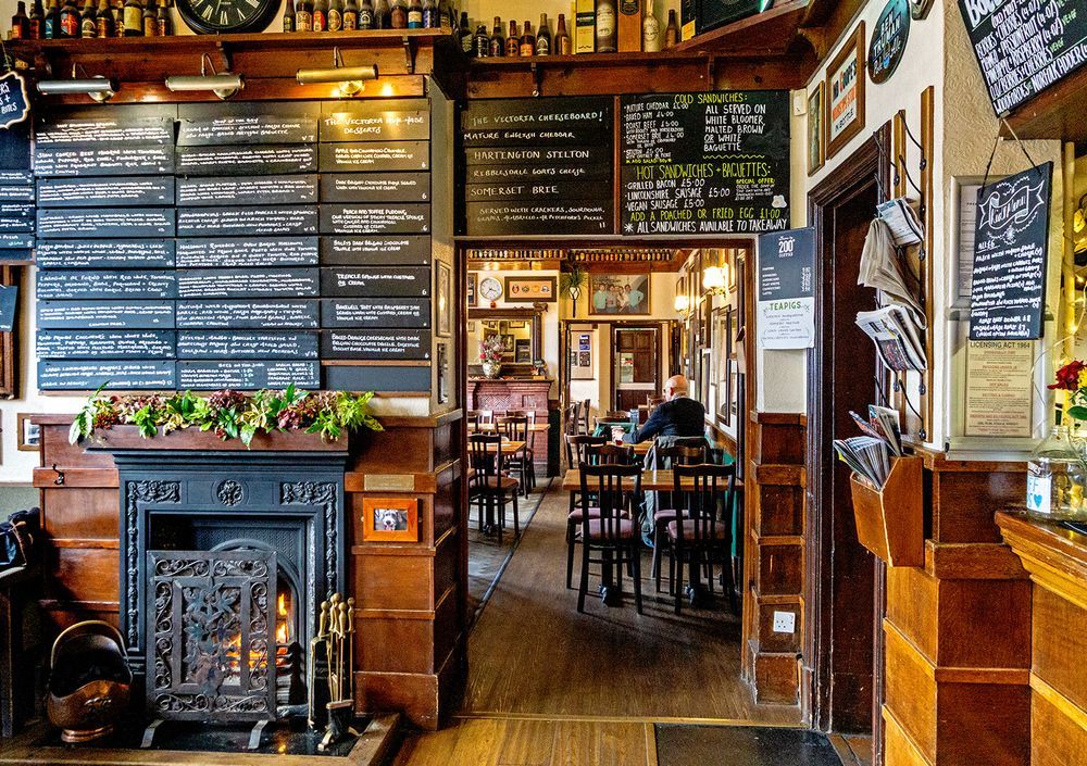 Main bar area with art deco styling on bar counter tand fireplace and view through to Smoke Room with multiple tables and chairs.