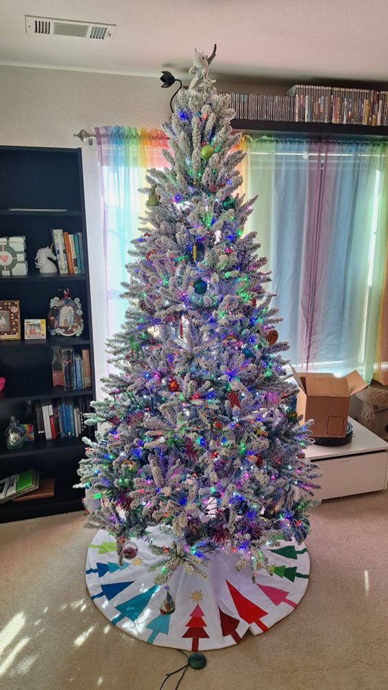 A christmas tree. The ornaments and light are multi-colored across the full spectrum. The tree skirt has rainbow trees on it. A plush dragon sits atop as a tree topper. A bookshelf is in the background 