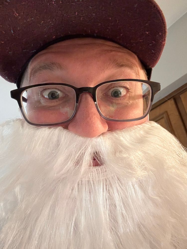 A guy in a red cap with glasses wearing a large white costume beard. He looks intense and proud of what he’s done. 