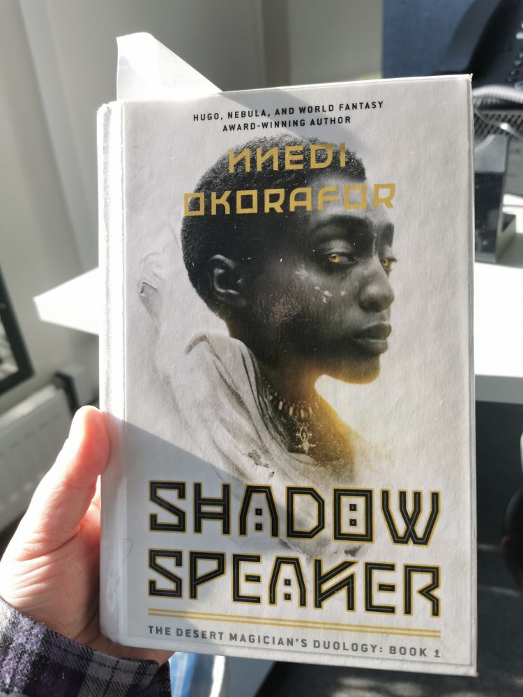 Forsiden til boka "shadow speaker" av nnedi okorafor. Den er hvit og har bilde av en mørkhudet jente med kort hår og gule øyne. 