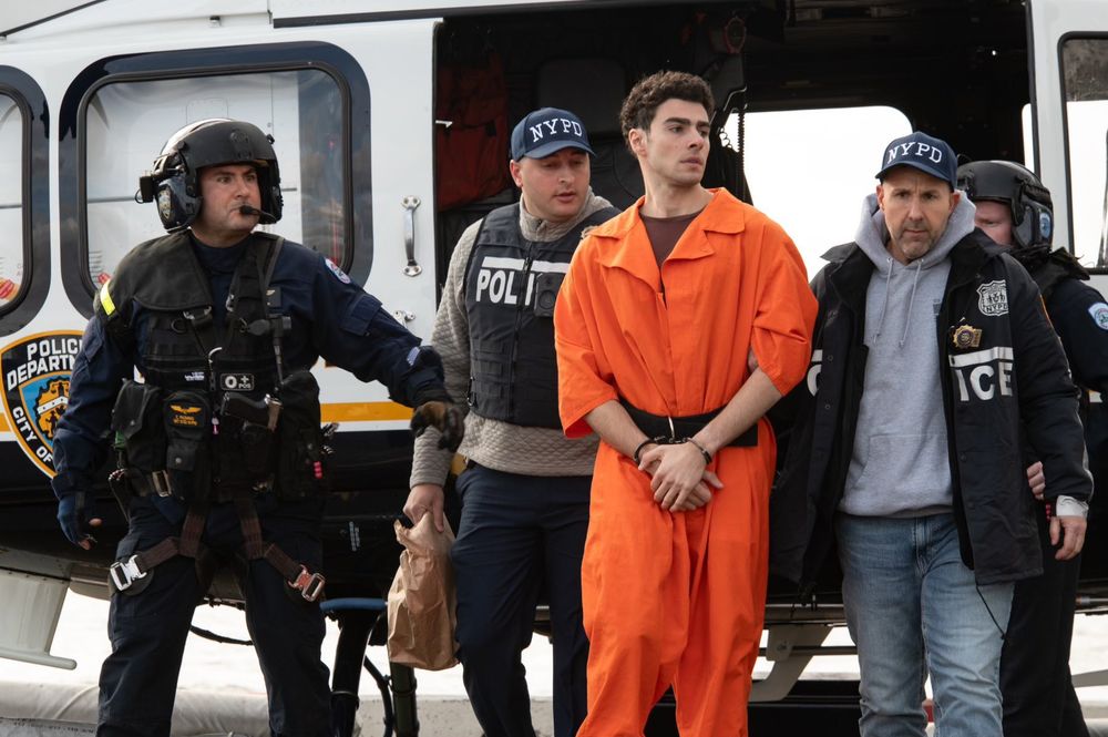 Luigi Mangione in orange jumpsuit in the center of the frame, flanked by law enforcement officers, his hands are clasped and bound in front of him he looks off to the side pensively, seriously does this guy just not photograph poorly at all? 