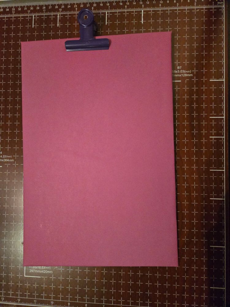 Auf einer karierten Unterlage liegt etwas rechteckiges Pinkfarbenes mit einer lila Klammer im oberen Bereich. 