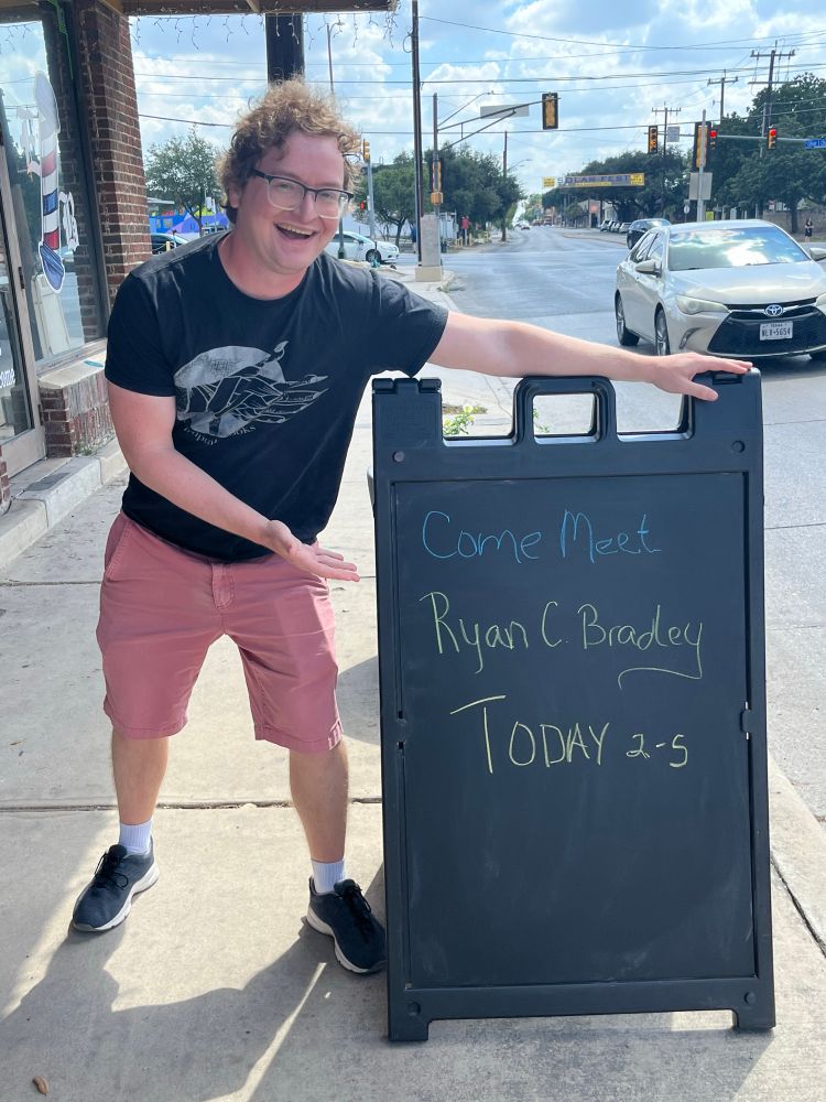 Ryan Bradley by a chalkboard sign advertising he’s signing from2-5 today 