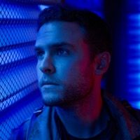 Leo Fitz from Agents of Shield gazes pensively through a metal screen to the left. Everything is so blue you'd think it was from a Michael Bay movie 