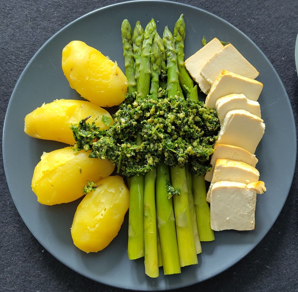 grauer Teller auf grauem Hintergrund, auf dem Teller befinden sich links 4 gepellte Kartoffeln, mittig grüne Spargelstangen getoppt mit grünem Pesto, rechts Räuchertofu in dicken Scheiben