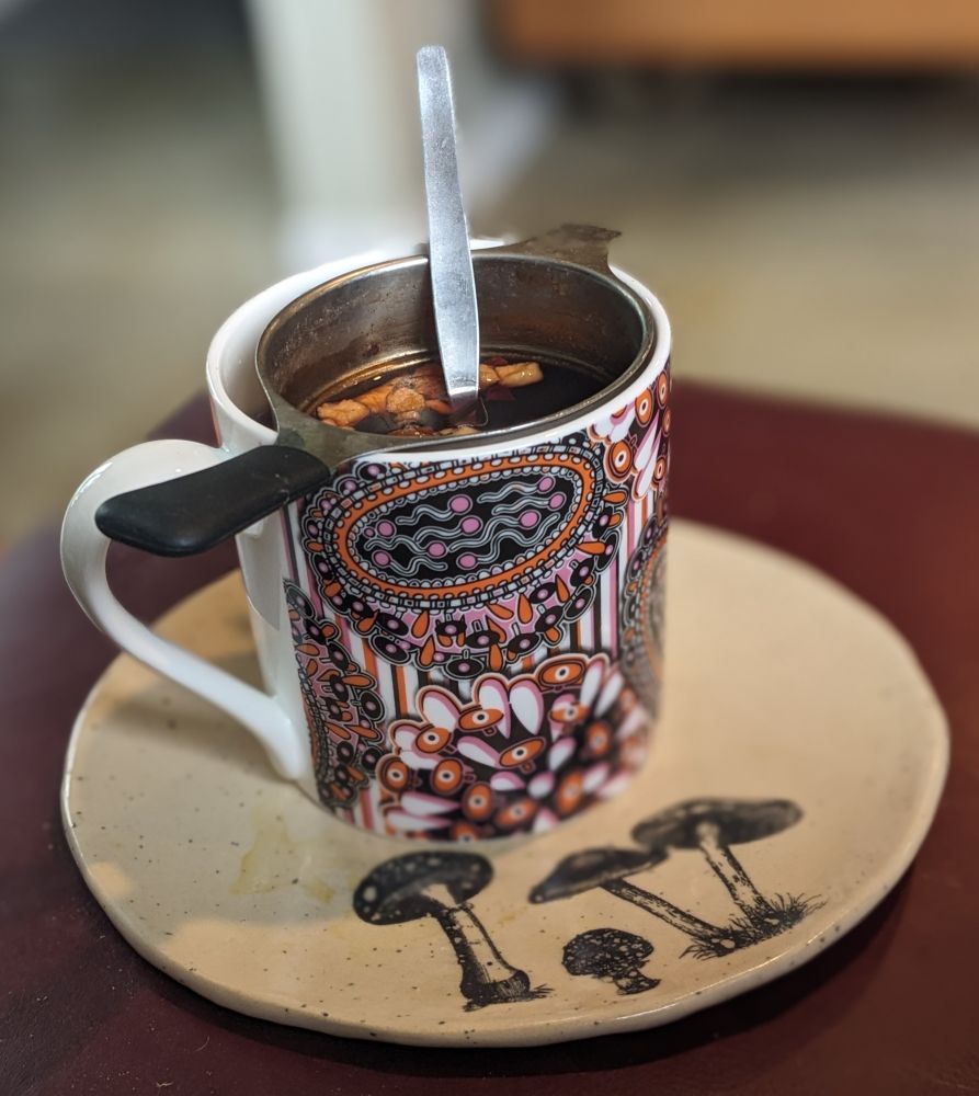 A colourful pink and orange mug with a tea strainer, and spoon sitting on a lovely cream coloured plate decorated with mushrooms 