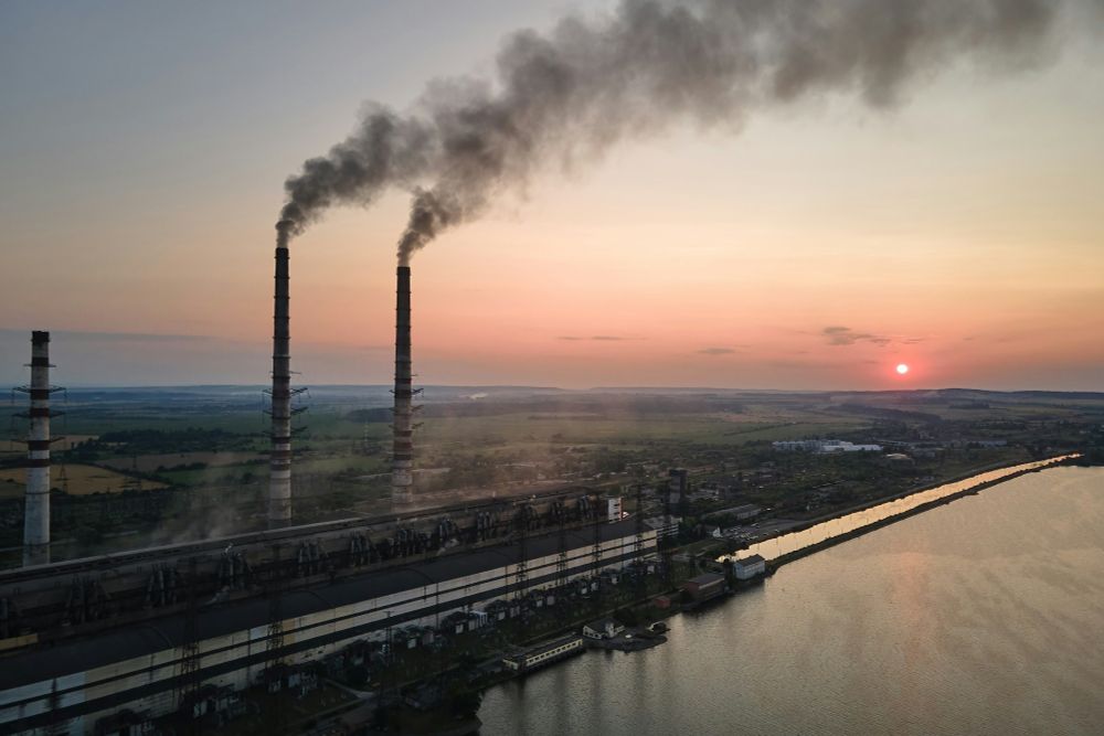 Aerial view of an industrial area with smokestacks emitting smoke beside a river at sunset. Photo credit: Getty Images via Unsplash.