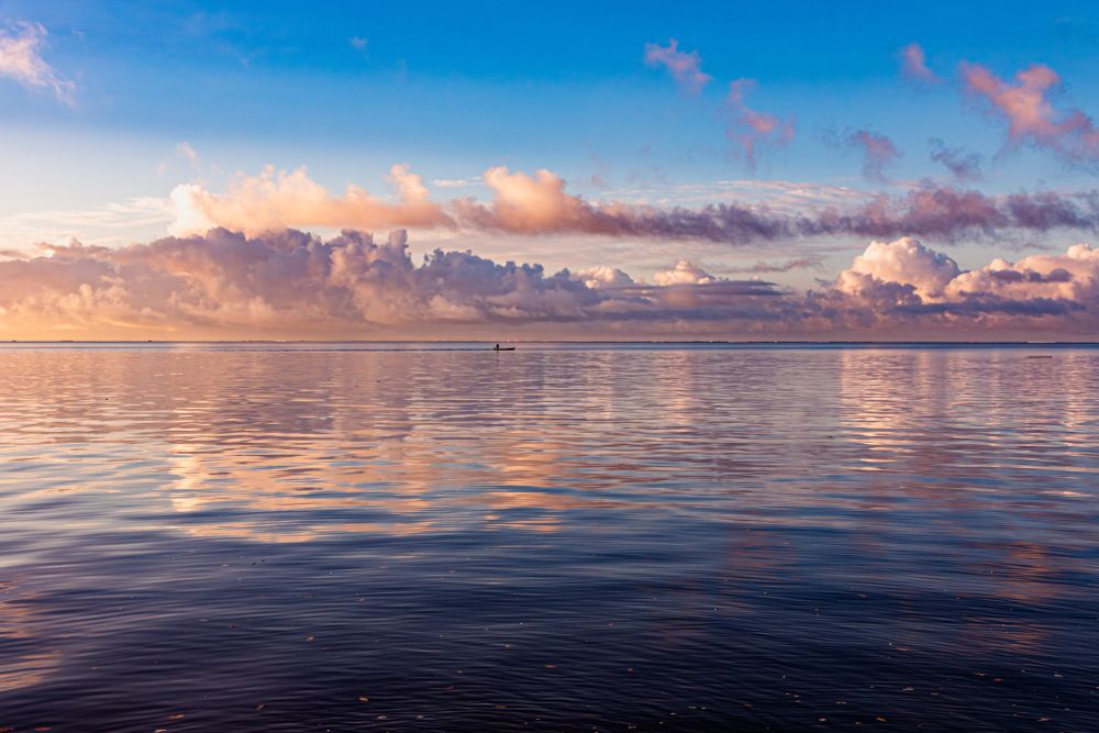 A serene image of a vast ocean under a colorful sky, with calm waters reflecting the light hues of the sky and a small boat visible in the distance. Photo credit: 2024 Weather Band Photo Contest submission "Sunrise at Suva Point" by Viniana Bau.
