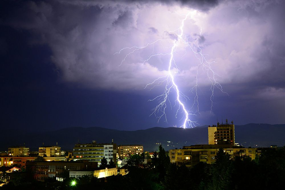 Nighttime view of a city skyline with lightning striking down behind the buildings under a stormy sky. Photo credit: Tsvetoslav Hristov via pexels.