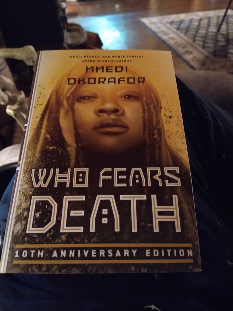 Cover of book: Who Fears Death written by Nnedi Okorafor. Book is in the foreground, resting on a person's knee, with a living room floor in the background.