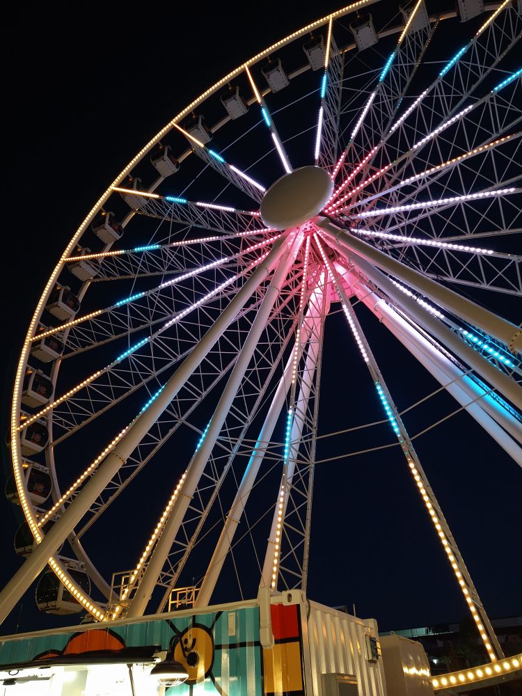 Large gondola style Ferris wheel with pink and blue lighting against dark purple dusk sky, in Kansas City Missouri 