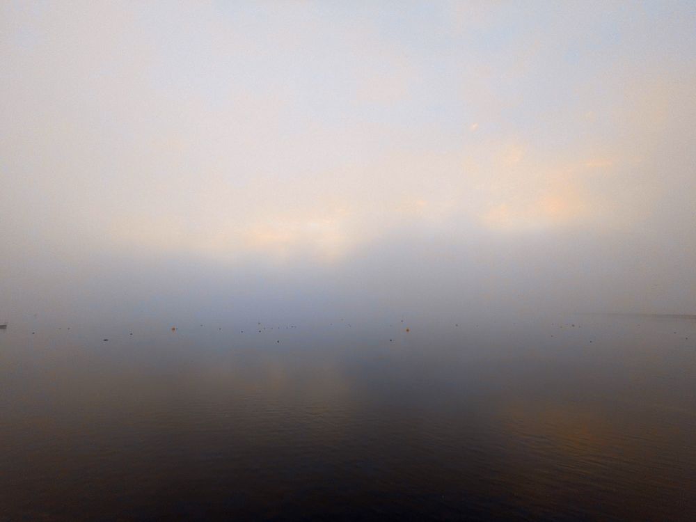 Icy frost over a reservoir, seen as swirling grey with hints of blue, yellow and orange. A few small dots of swimming birds can be seen in the lower portion of the picture, helping identify the water from the sky
